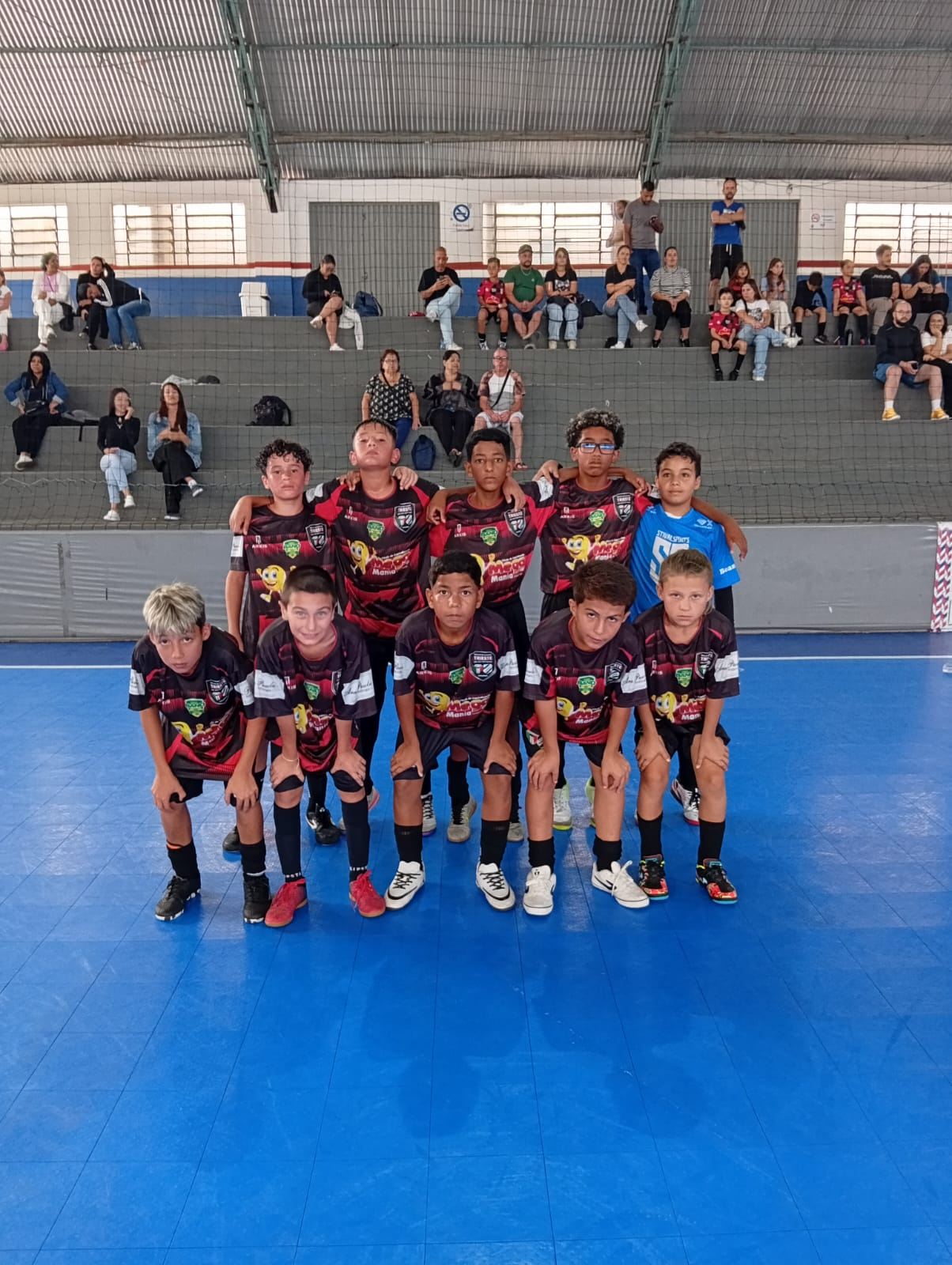 Equipe do Trieste posada na quadra azul de um ginásio, com torcida na arquibancada